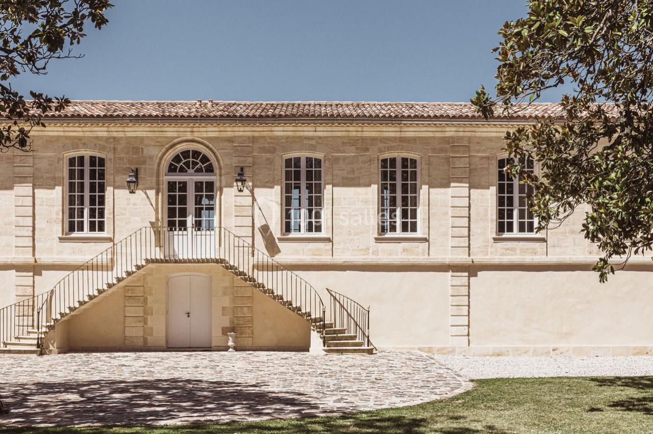 Façade d'un bâtiment en pierre avec escalier central, grandes fenêtres et cour pavée, entouré de verdure.
