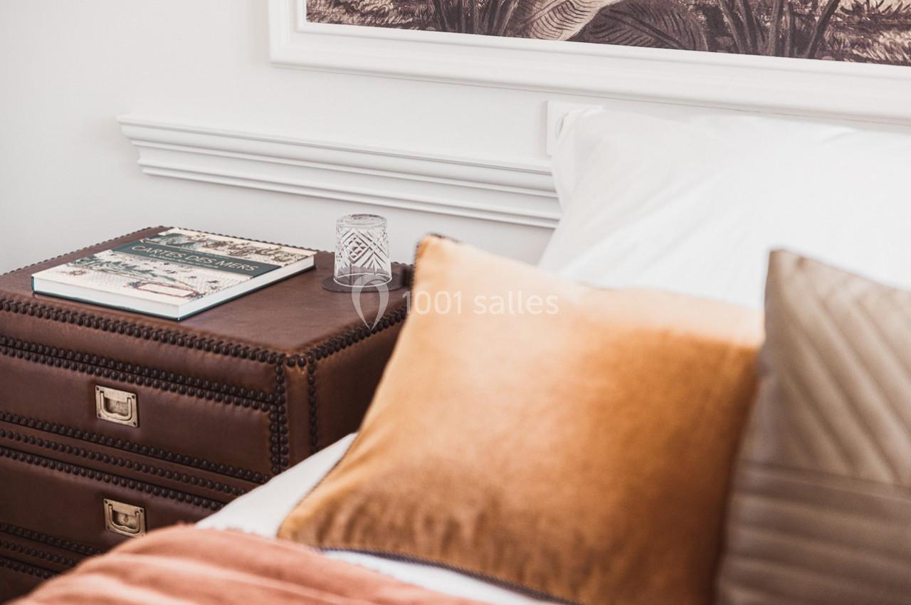 Table de chevet en cuir avec un livre, un verre et un lit avec des coussins dans une chambre lumineuse.