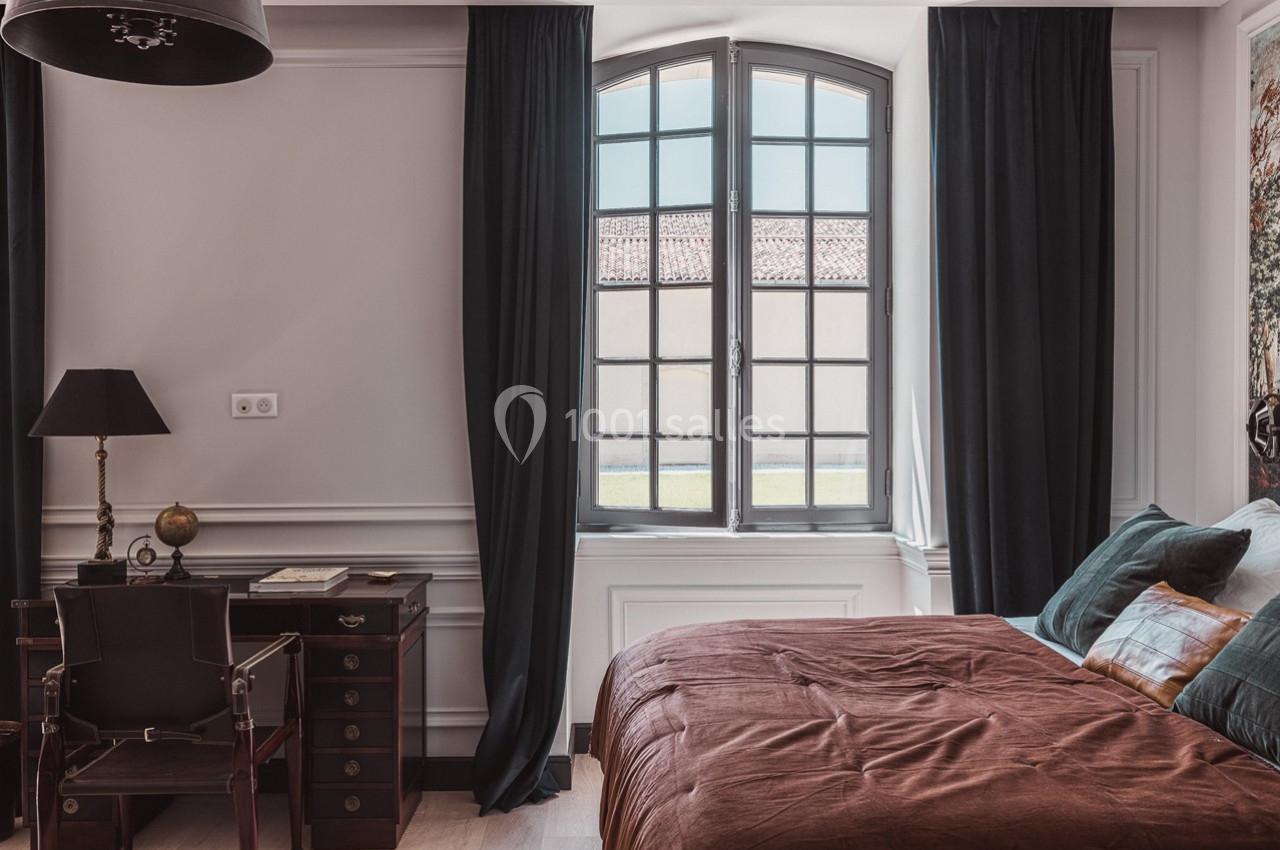 Chambre lumineuse avec un lit, un bureau en bois, une lampe et une grande fenêtre encadrée de rideaux sombres.