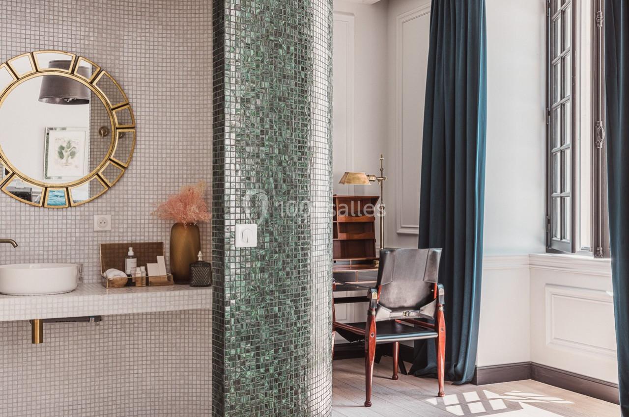 Salle de bain lumineuse avec miroir rond doré, vasque blanche, cloison en mosaïque verte et bureau en arrière-plan.