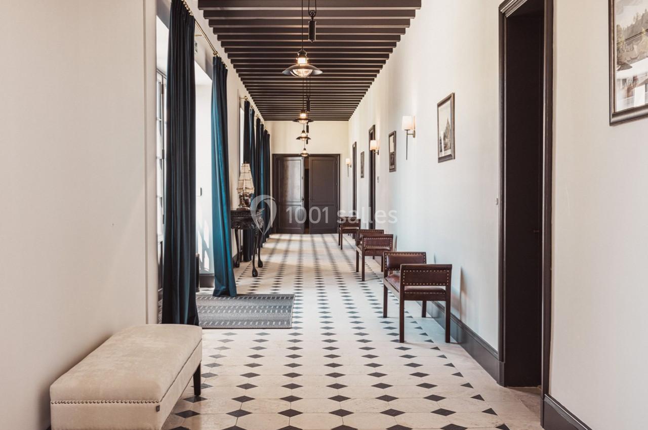 Couloir lumineux avec sol en carrelage noir et blanc, bancs en bois, rideaux sombres et éclairage suspendu.
