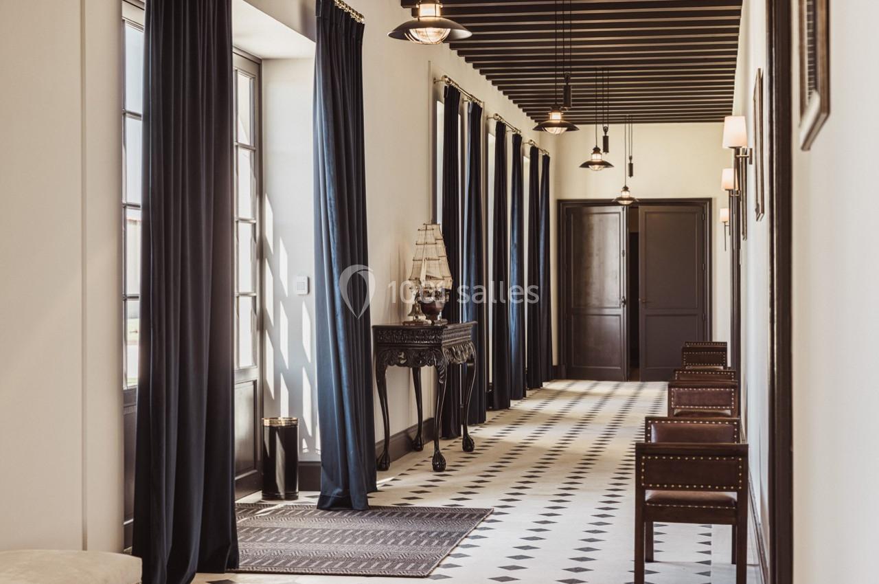 Couloir lumineux avec sol en damier, rideaux sombres, meubles en bois et maquette de bateau sur une console.