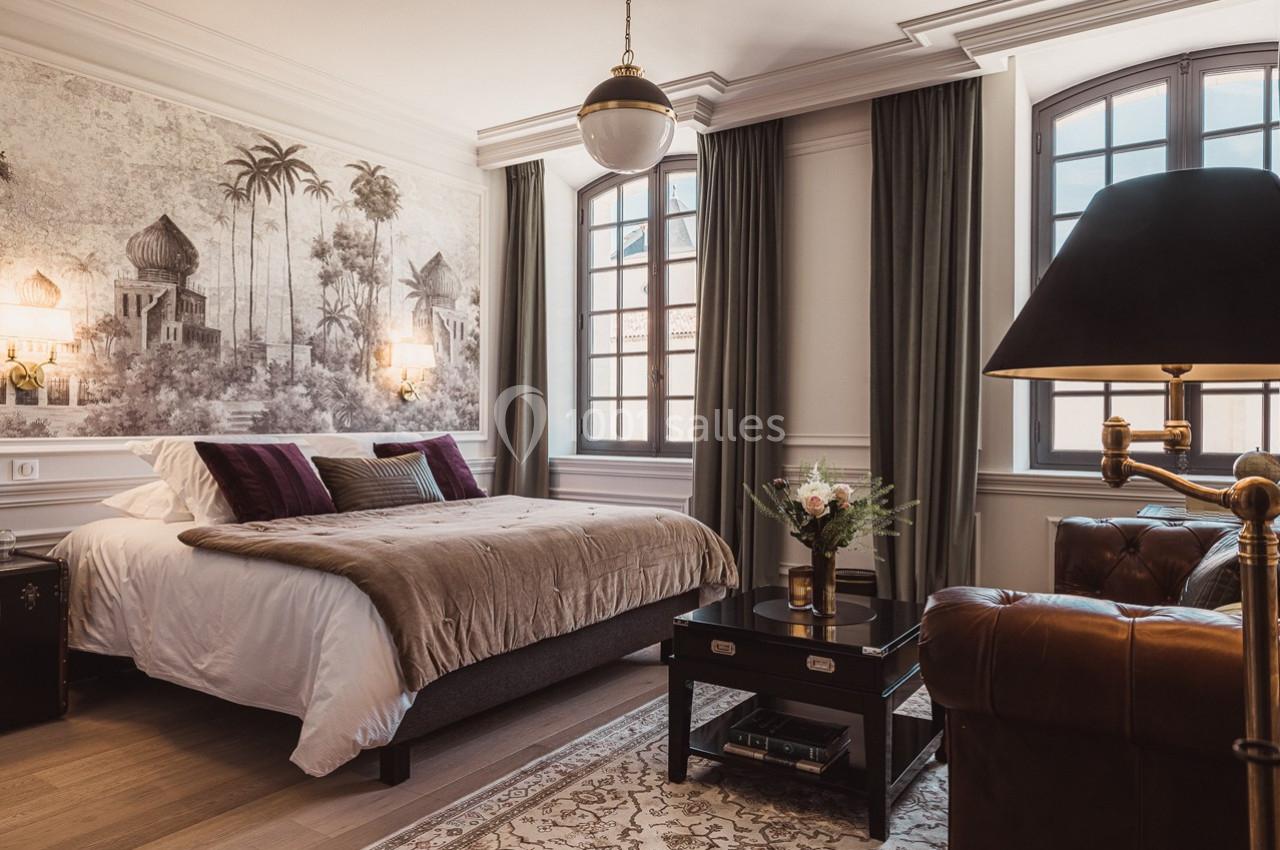 Chambre élégante avec lit double, décoration murale exotique, grandes fenêtres, fauteuil en cuir et table basse fleurie.