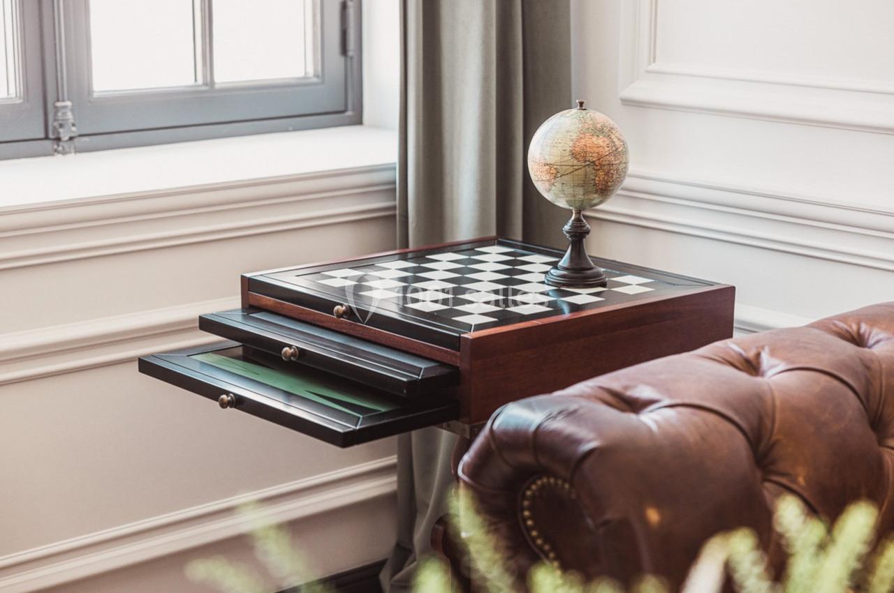 Table d'échecs en bois avec tiroirs ouverts, globe terrestre en décoration, près d'un canapé en cuir et d'une fenêtre.