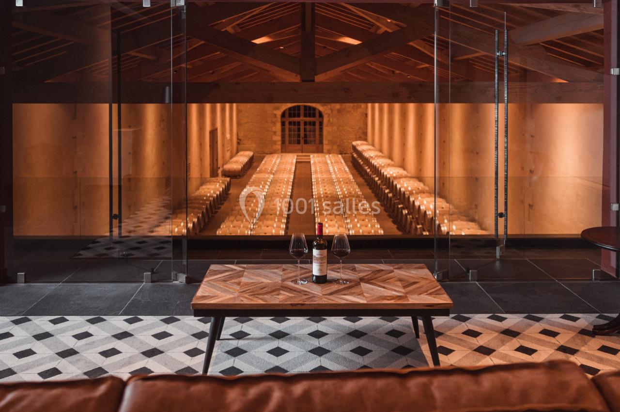 Vue d'une cave à vin avec des rangées de tonneaux, observée depuis un salon avec une table et un verre de vin.