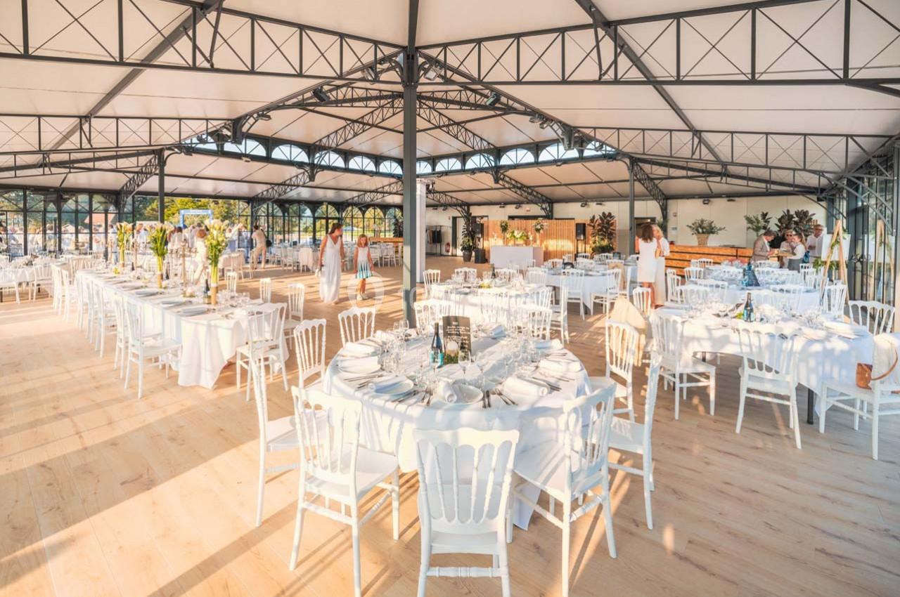 Salle de réception lumineuse avec tables rondes dressées, chaises blanches et grande structure métallique apparente.