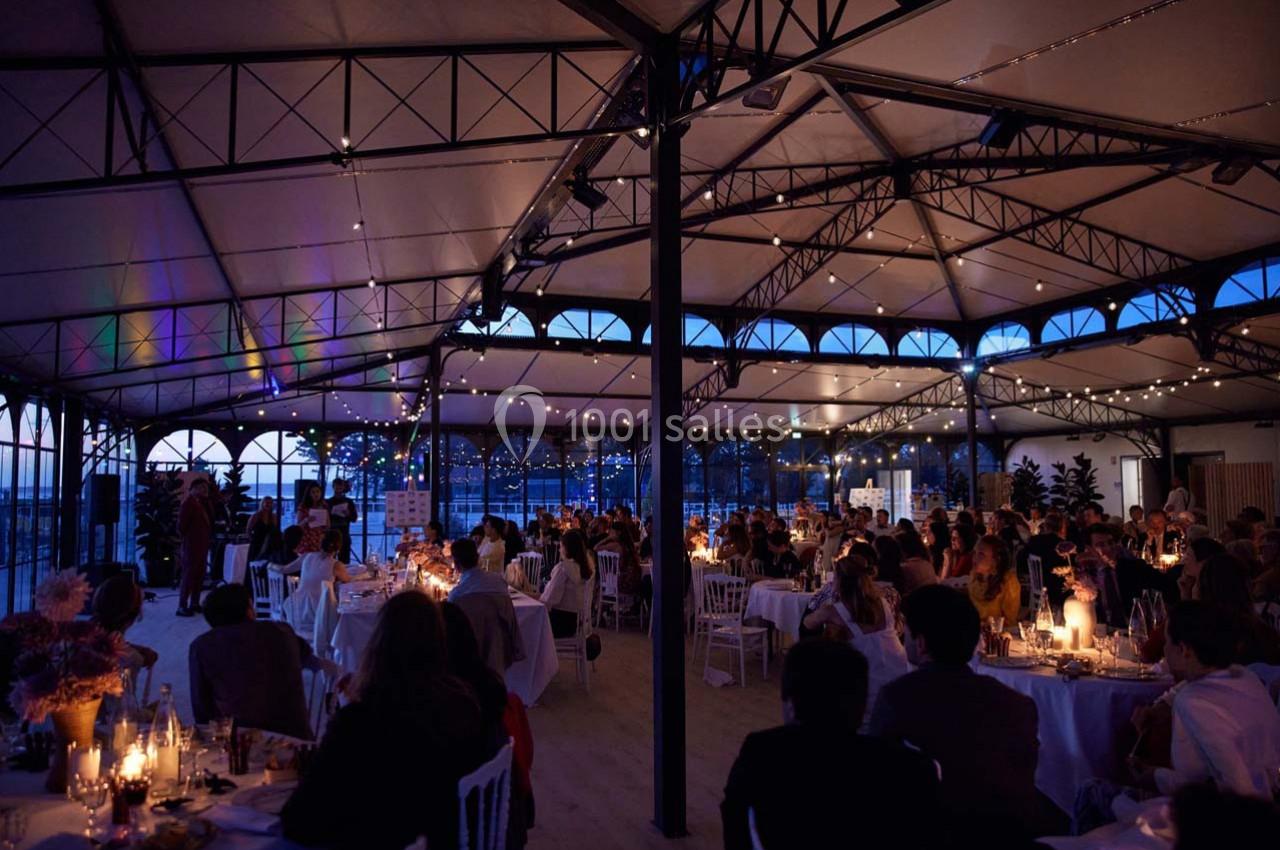 Salle de réception illuminée en soirée, avec des invités assis à des tables décorées et des bougies allumées.