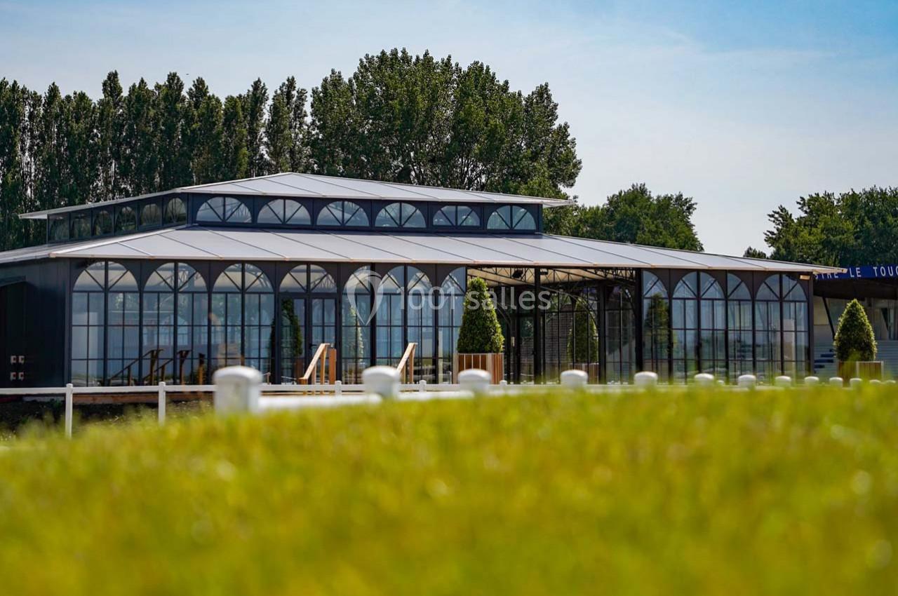 Bâtiment moderne avec une structure en verre et métal entouré de verdure, sous un ciel dégagé.