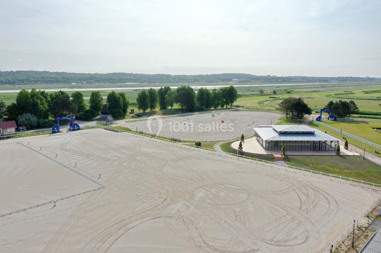 Vue d'un grand terrain équestre en sable avec des espaces délimités, entouré de verdure et d'un bâtiment moderne.