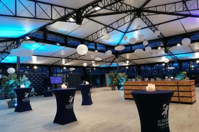 Bâtiment vitré éclairé en soirée, entouré d'arbres et d'une clôture blanche, avec quelques personnes à l'extérieur.