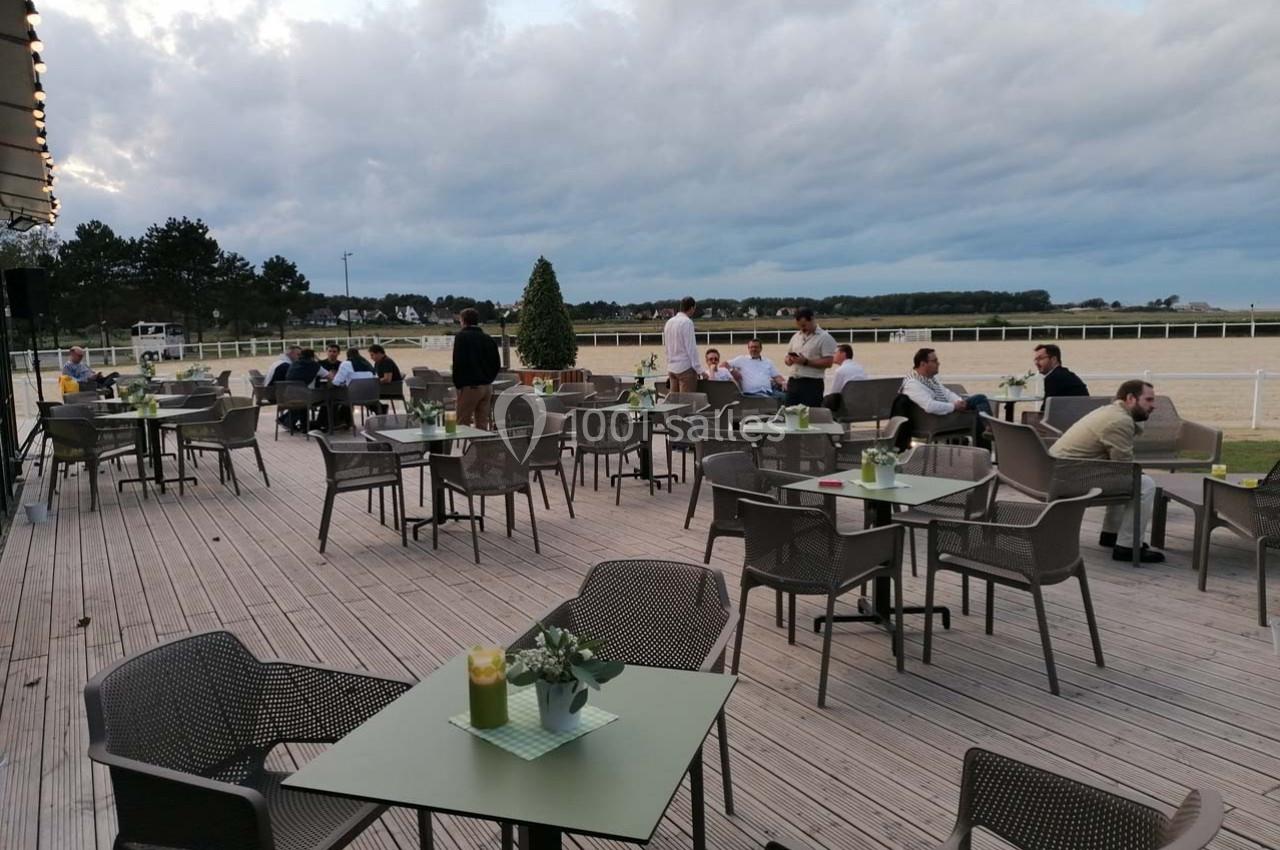 Terrasse en bois avec tables et chaises occupées par des personnes, vue sur une piste et un paysage arboré.