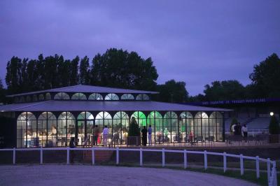 Bâtiment vitré éclairé en soirée, entouré d'arbres et d'une clôture blanche, avec quelques personnes à l'extérieur.