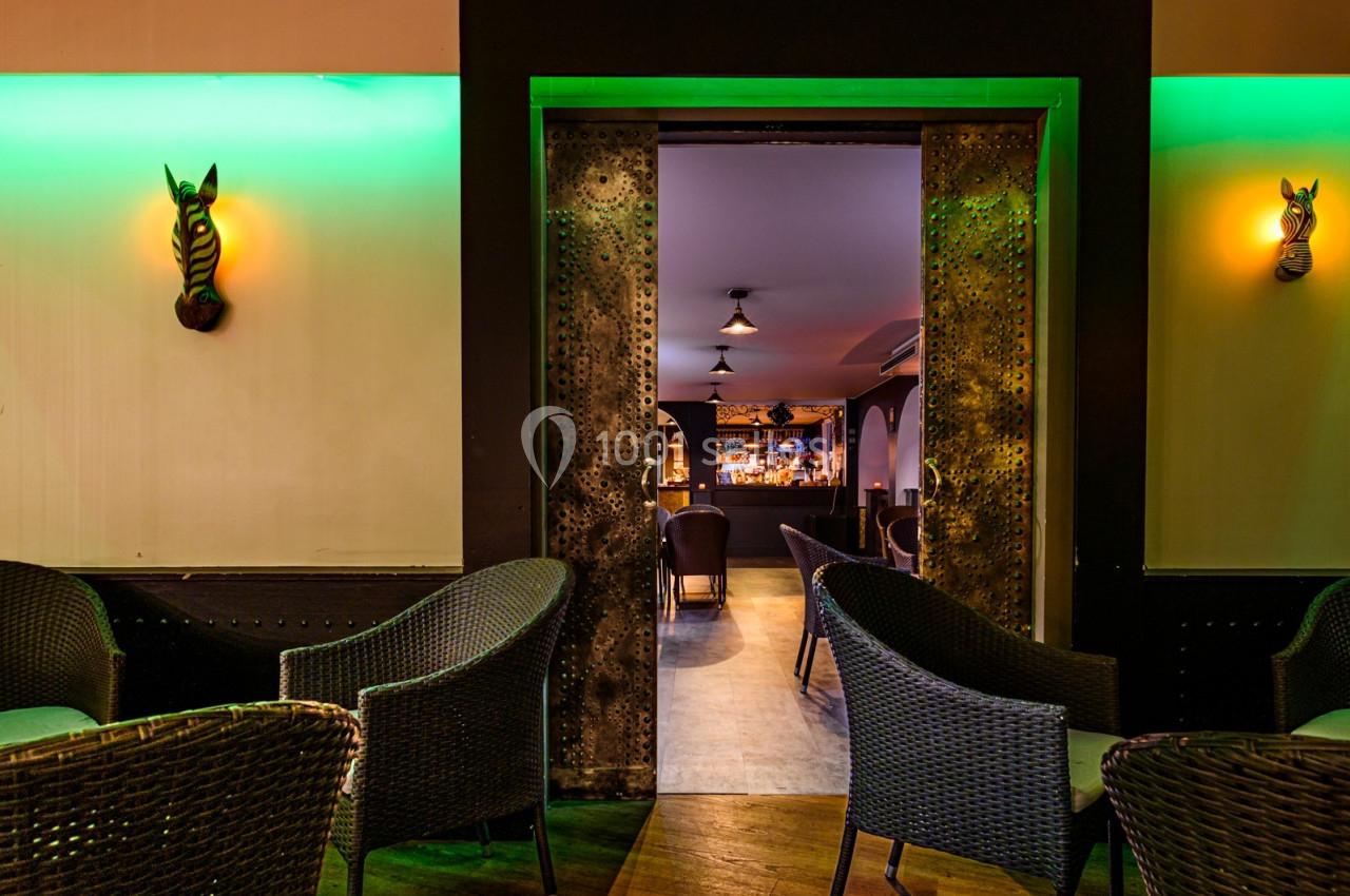 Vue d'un intérieur de bar avec des fauteuils en osier, une porte ouverte et un éclairage tamisé vert et jaune.