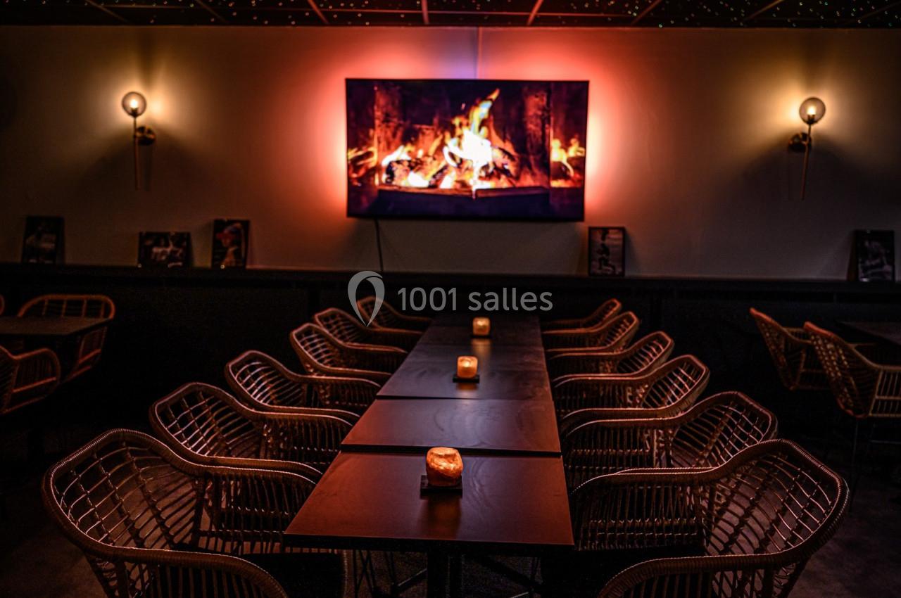 Salle sombre avec des tables alignées, chaises en rotin et un écran affichant une cheminée enflammée sur le mur.