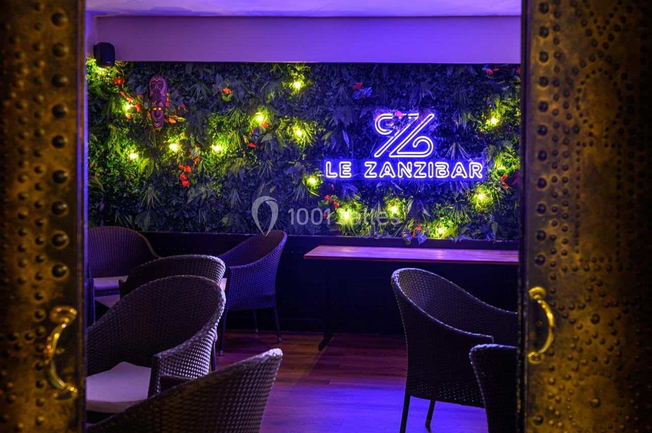 Mur végétal éclairé avec un néon ’Le Zanzibar’, entouré de fauteuils en rotin et tables en bois dans un espace intérieur.