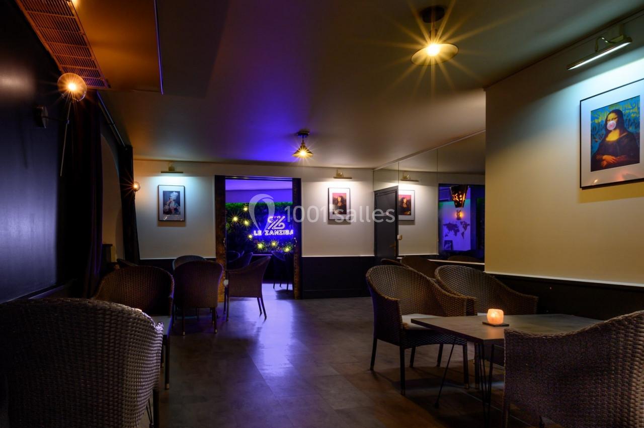 Salle de bar avec fauteuils en osier, éclairage tamisé et enseigne lumineuse visible au fond.