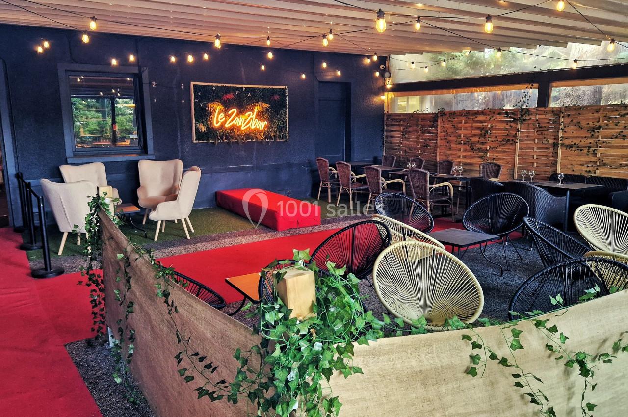 Espace extérieur aménagé avec fauteuils, tables, guirlandes lumineuses et panneau lumineux sur un mur sombre.