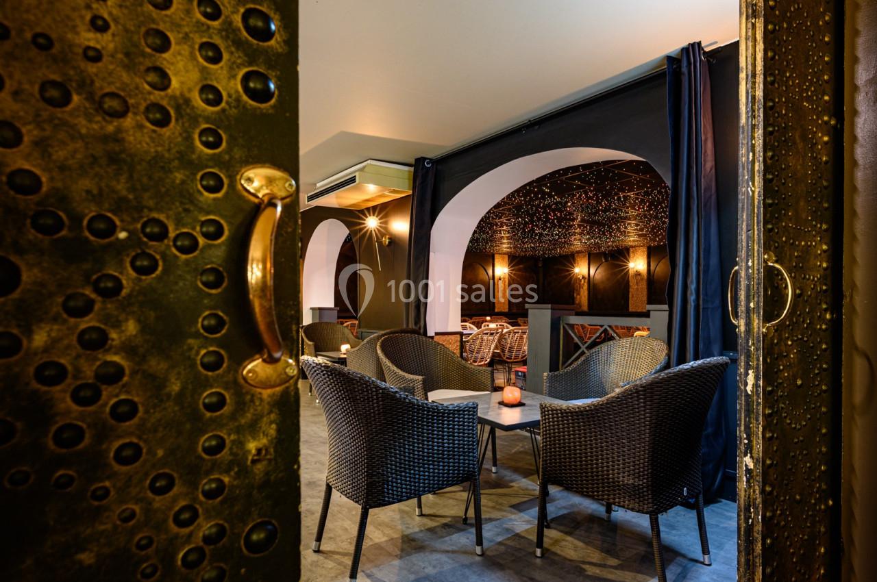 Salle cosy avec fauteuils en rotin, éclairage tamisé et décorations murales sombres, vue depuis une porte dorée.