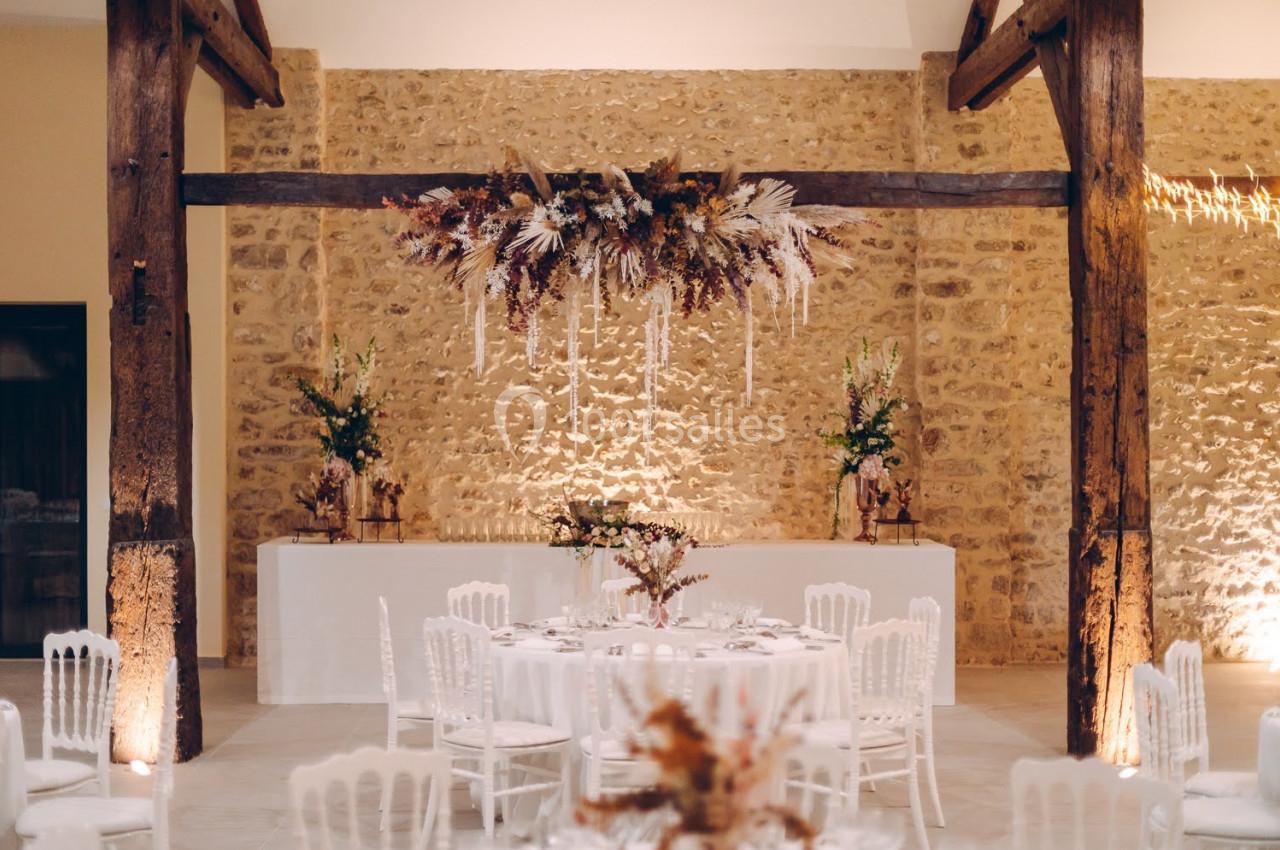 Salle de réception décorée avec des tables rondes, des chaises blanches et une suspension florale au plafond.