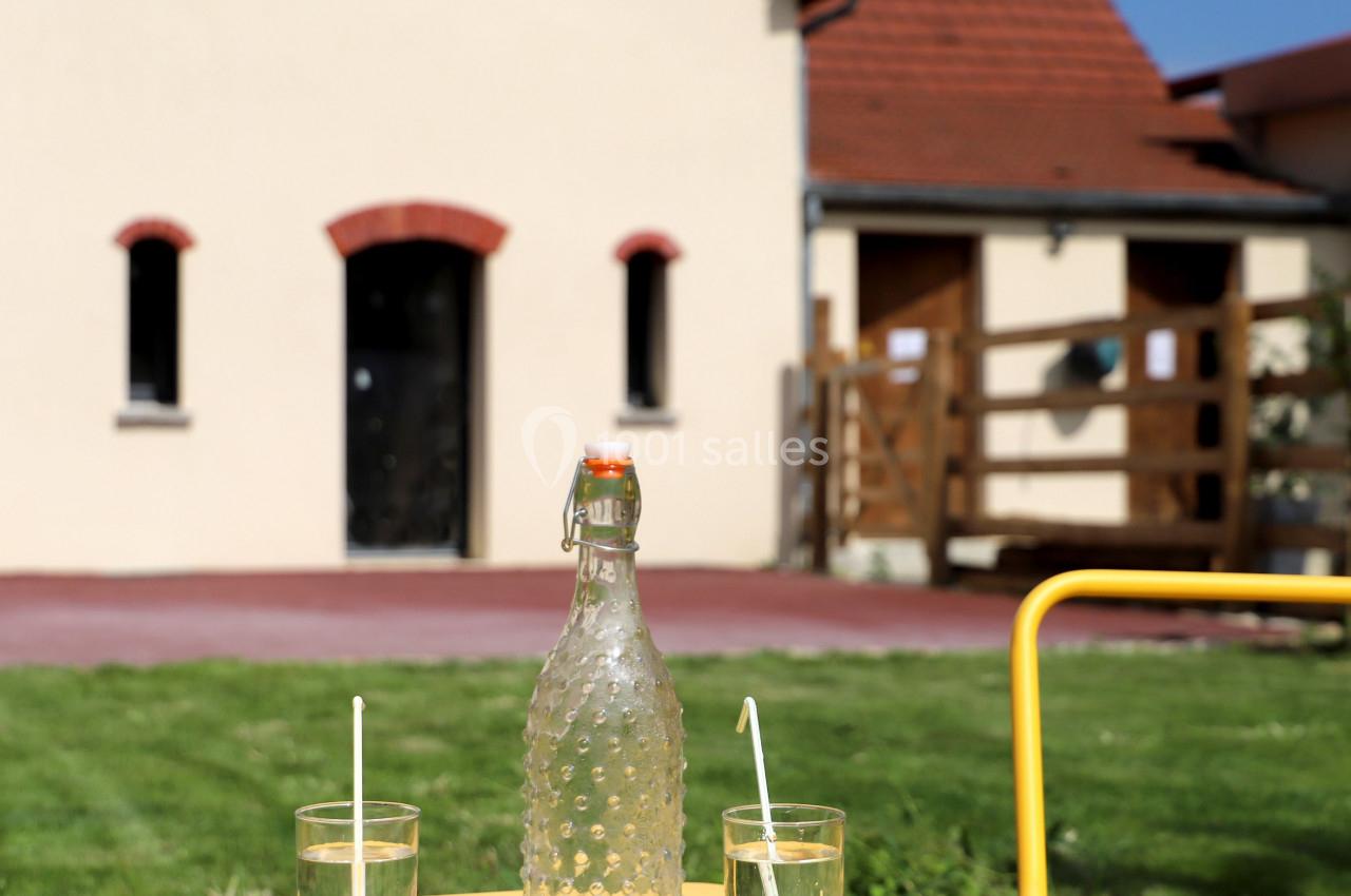 Une bouteille en verre et deux verres d'eau avec pailles sur une table jaune, devant une maison avec jardin.