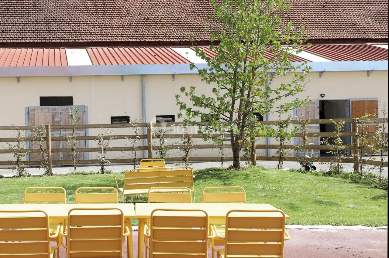Des tables et chaises jaunes sont disposées sur une terrasse en plein air, près d'un arbre et d'un bâtiment clair.