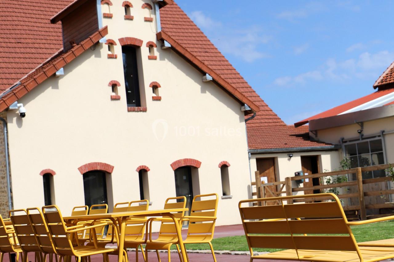 Façade d'un bâtiment à toit rouge avec une terrasse équipée de tables et chaises jaunes sur sol rouge.