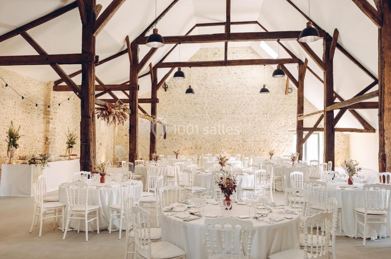Salle de réception lumineuse avec des tables rondes dressées, des chaises blanches et une charpente en bois apparente.