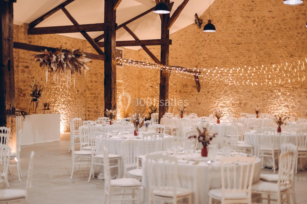 Salle de réception décorée avec des tables rondes, chaises blanches, guirlandes lumineuses et murs en pierre apparente.