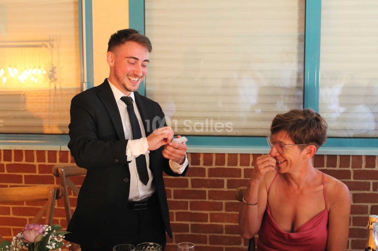 Un homme en costume réalise un tour de magie devant une femme souriante assise à une table.