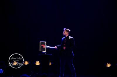 Un magicien sur scène tenant une baguette devant un accessoire éclairé dans un décor sombre.