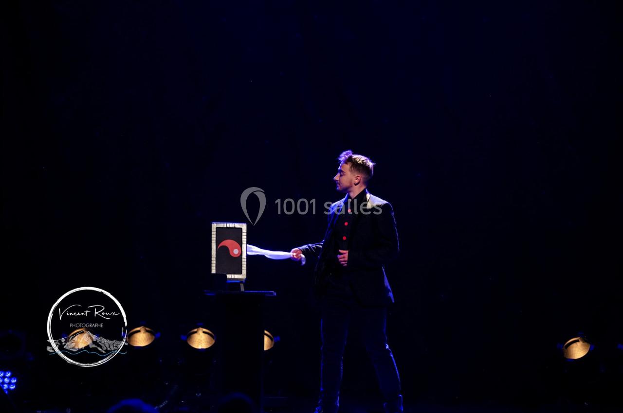 Un magicien sur scène tenant une baguette devant un accessoire éclairé dans un décor sombre.