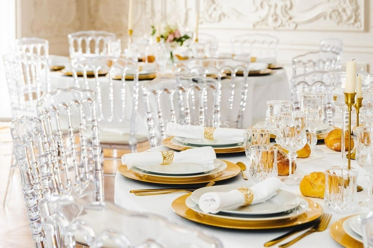 Tables élégamment dressées avec nappes blanches, assiettes dorées, serviettes roulées et chaises transparentes.