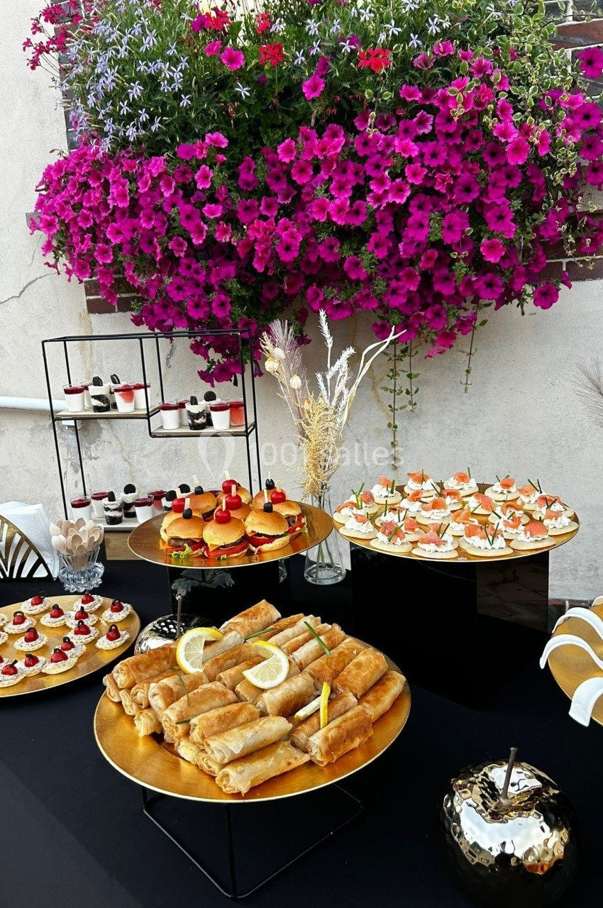 Buffet extérieur avec assortiment de bouchées salées et sucrées, décoré de fleurs violettes en arrière-plan.
