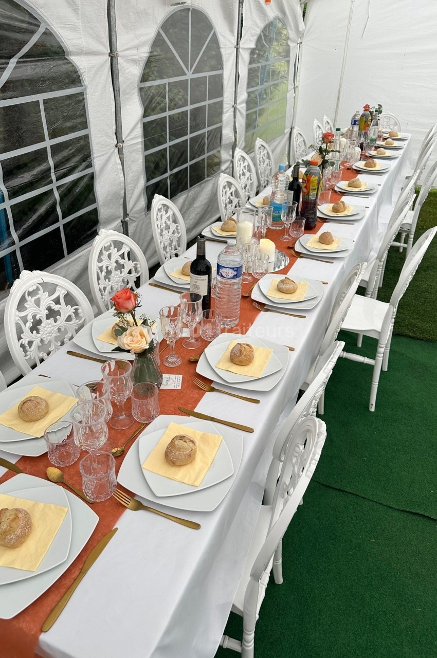 Table dressée pour un repas festif sous une tente, avec nappes orange, assiettes, verres, pains et décorations florales.