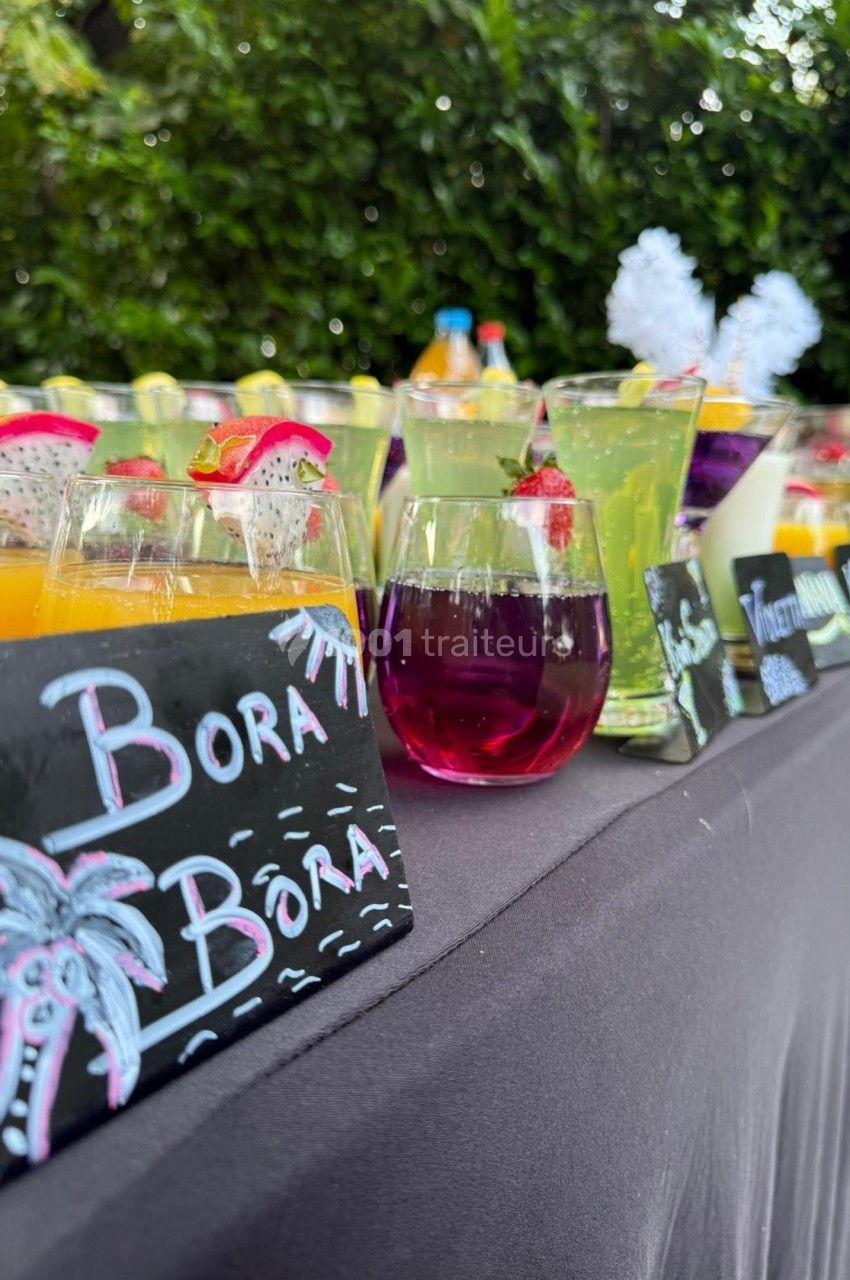 Table avec des cocktails colorés et étiquettes écrites à la craie, disposée en extérieur avec un fond de verdure.