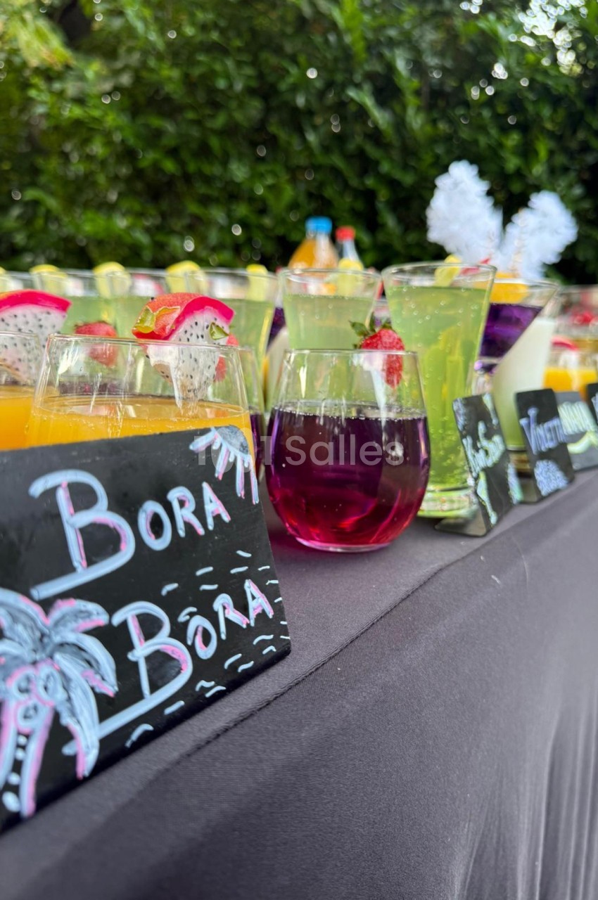 Table avec des cocktails colorés et étiquettes écrites à la craie, disposée en extérieur avec un fond de verdure.