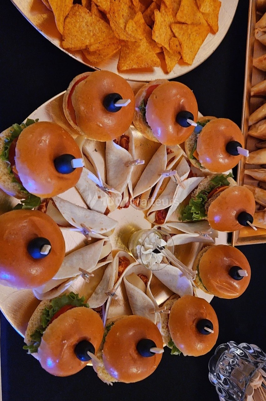 Plateau de mini-burgers garnis et wraps disposés en cercle, entourés de chips triangulaires sur une table noire.