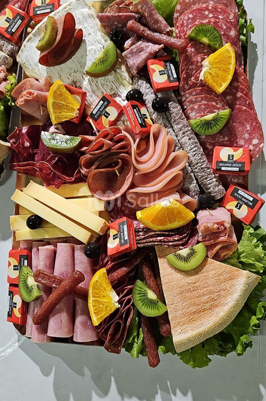 Plateau de charcuterie et fromages avec fruits frais, olives et tranches de pain, présenté sur un lit de salade verte.