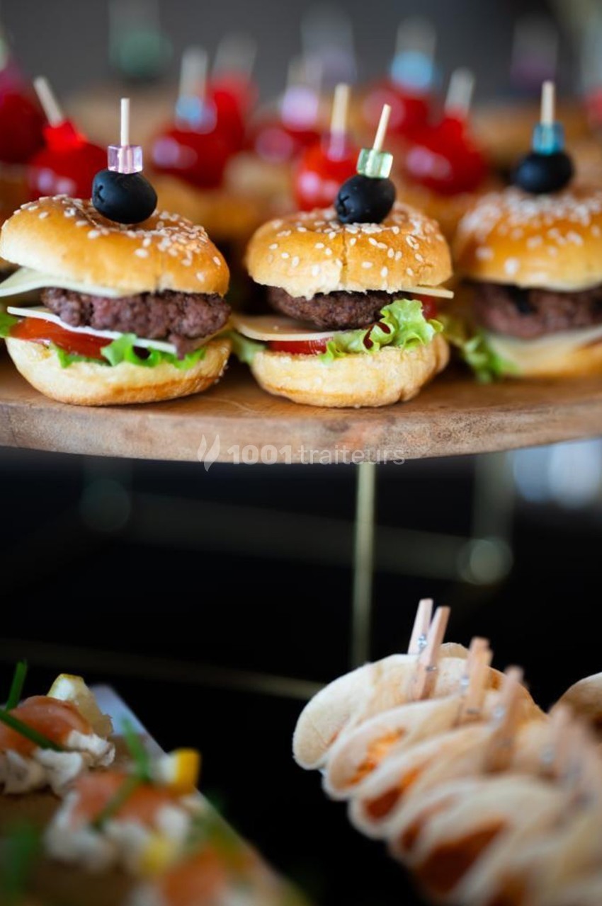 Mini burgers garnis de salade, tomate et fromage sur un plateau en bois, avec d'autres amuse-bouches en arrière-plan.