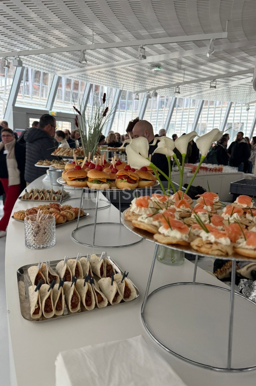Buffet avec plateaux de canapés, mini-burgers, tacos et fleurs blanches, dans une salle lumineuse avec des convives.