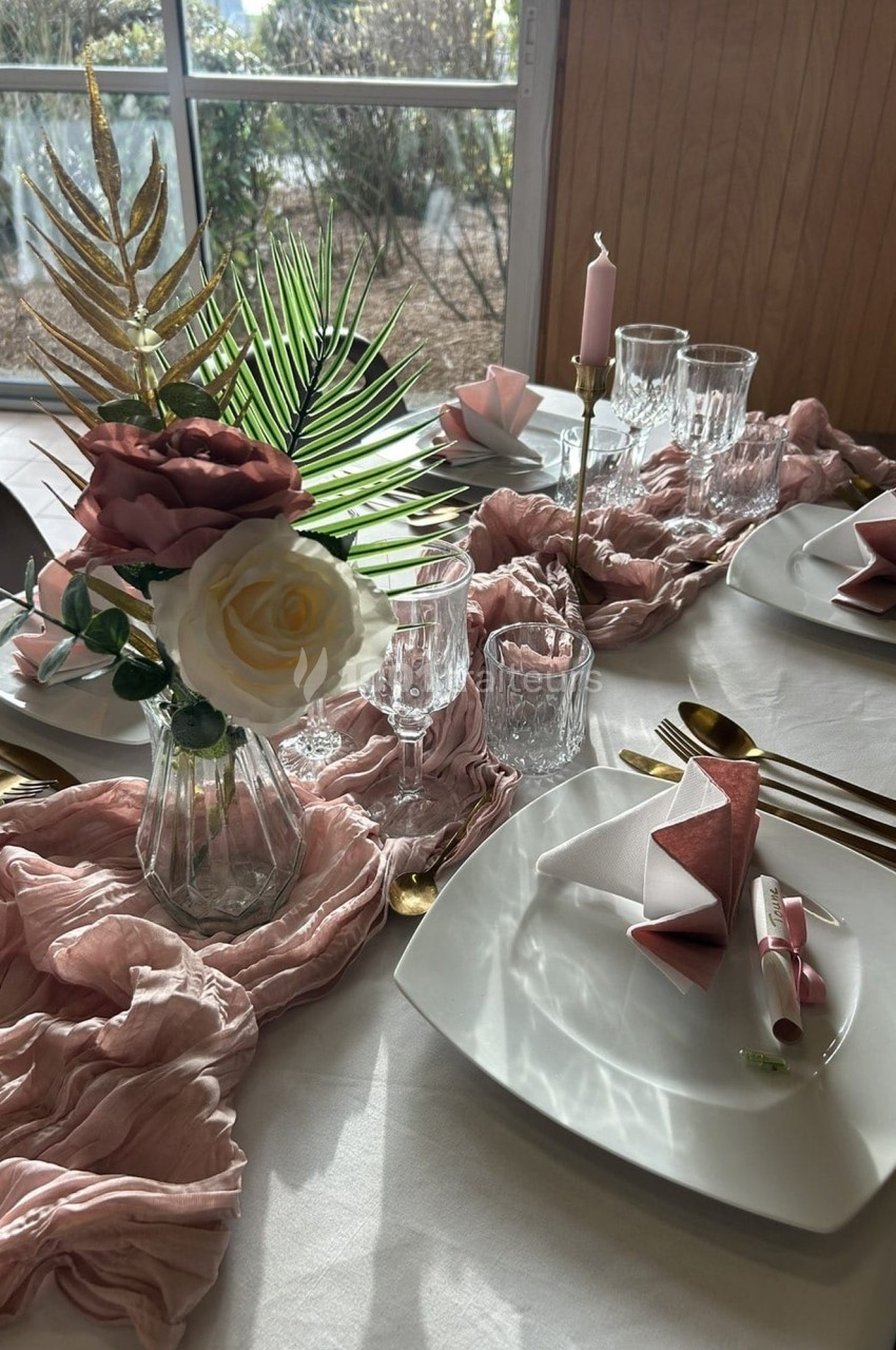 Table élégamment dressée avec des assiettes blanches, serviettes roses pliées, verres, bougies et décoration florale.