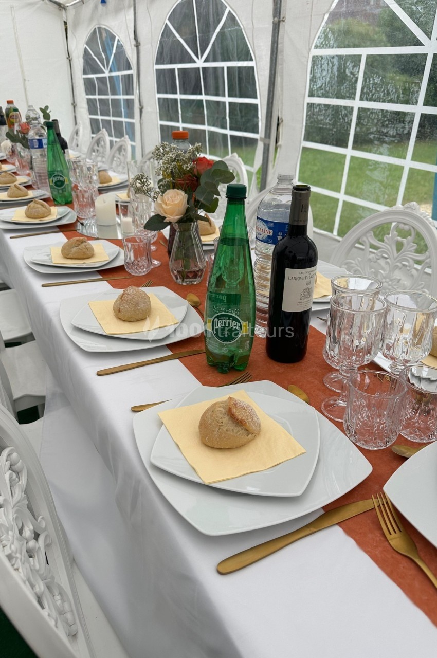 Table dressée sous une tente avec assiettes, verres, bouteilles d'eau et de vin, pain et décoration florale.