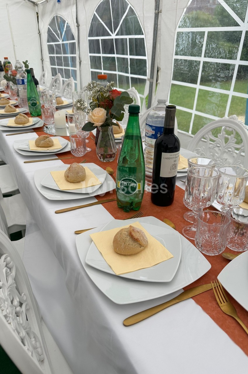 Table dressée sous une tente avec assiettes, verres, bouteilles d'eau et de vin, pain et décoration florale.
