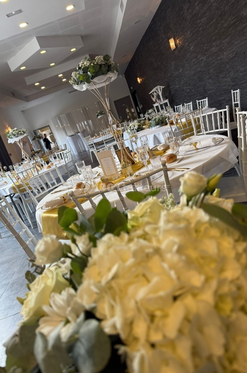 Salle de réception élégamment décorée avec des tables dressées, des chaises blanches et des compositions florales blanches.