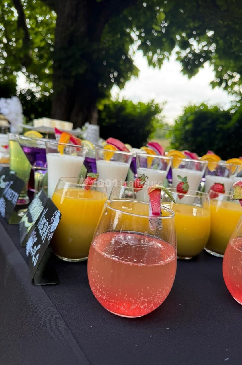 Verres de cocktails colorés disposés sur une table noire en extérieur, entourés de verdure.