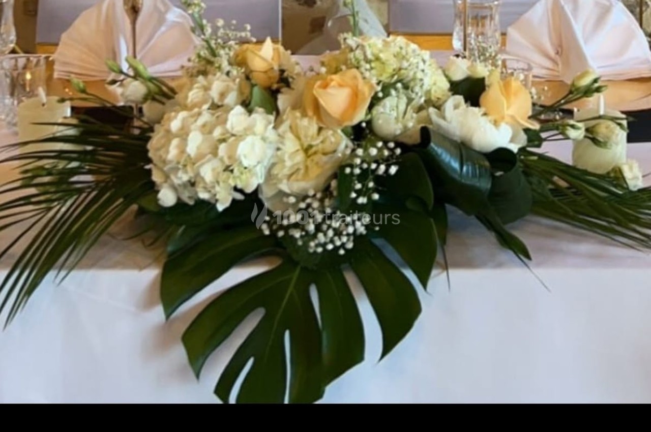 Arrangement floral avec roses jaunes, hortensias blancs et feuillages tropicaux, posé sur une table blanche.