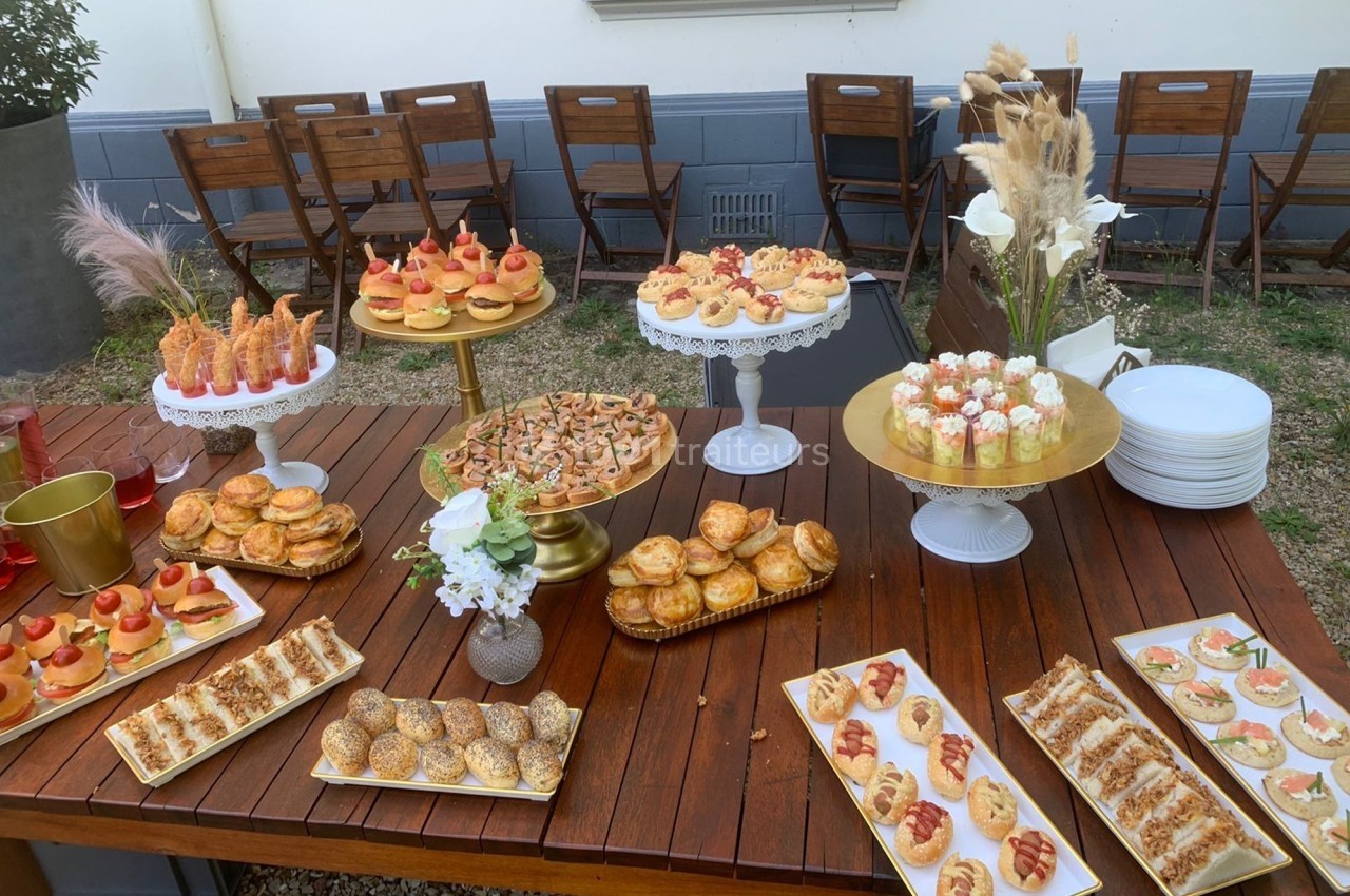 Table en bois garnie de divers amuse-bouches et desserts, disposés sur des plateaux et présentoirs dans un jardin.