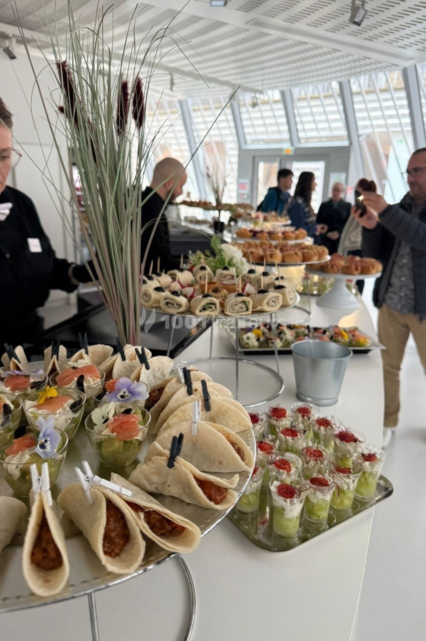 Buffet avec wraps, verrines et plats variés, présenté dans un espace lumineux avec des convives en arrière-plan.