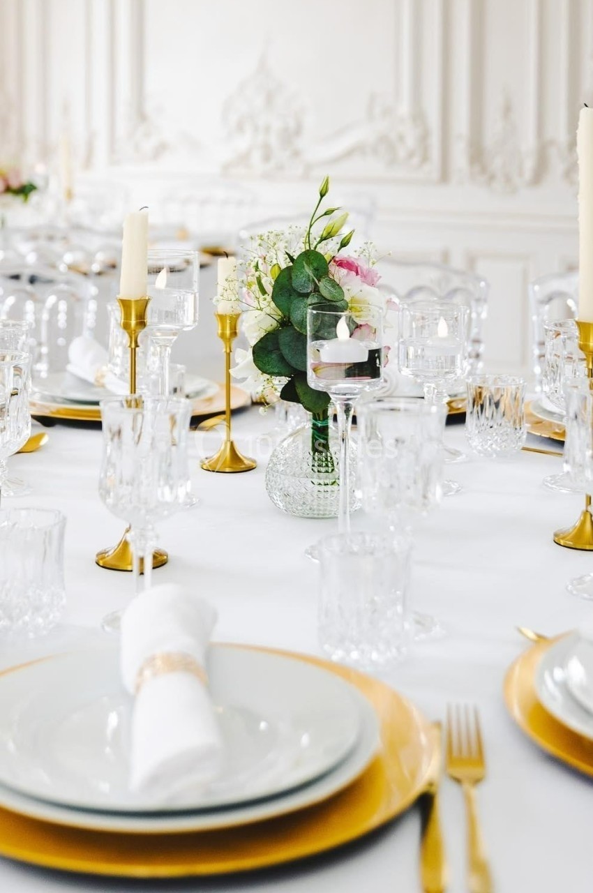 Table élégamment dressée avec vaisselle blanche, sous-assiettes dorées, chandeliers et bouquet floral central.