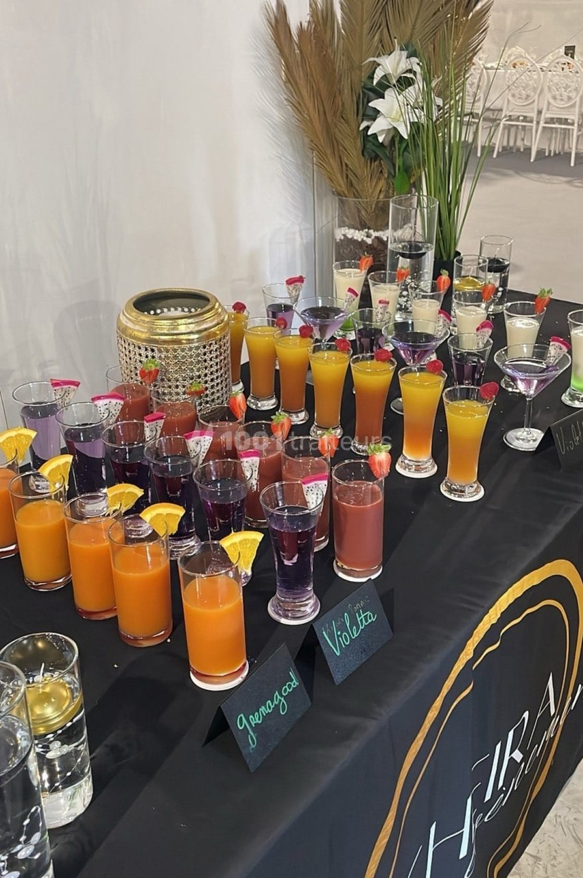 Table garnie de cocktails colorés avec des étiquettes indiquant les saveurs, décorée de plantes et bougies.